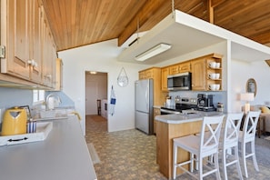 Una cocina con armarios de madera, microondas, refrigerador y un comedor con sillas blancas.