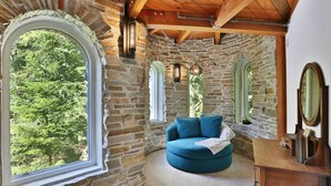Ein Zimmer mit Steinwänden, einem runden, blauen Sofa, Holzbalken und einem großen Fenster mit Blick auf Grünflächen.