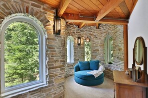 A stone-walled room with a round blue sofa, wooden beams, and a large window overlooking greenery. - Wiseman Castle- Beautiful Waterfalls! (Sedro-Woolley)