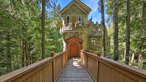 Eine Holzbrücke führt zu einem Steinhaus mit Balkon und Flaggen, umgeben von dichtem Wald.