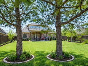 Un patio trasero residencial con una casa, dos árboles grandes, un patio y un césped bien cuidado.