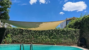 Una piscina con una vela de sombra, rodeada de exuberante vegetación y un edificio al fondo.