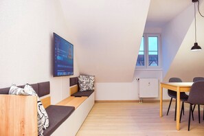 A modern loft bedroom with a loft bed, a dining area with a table and chairs, a large TV mounted on the wall, and a window providing natural light.