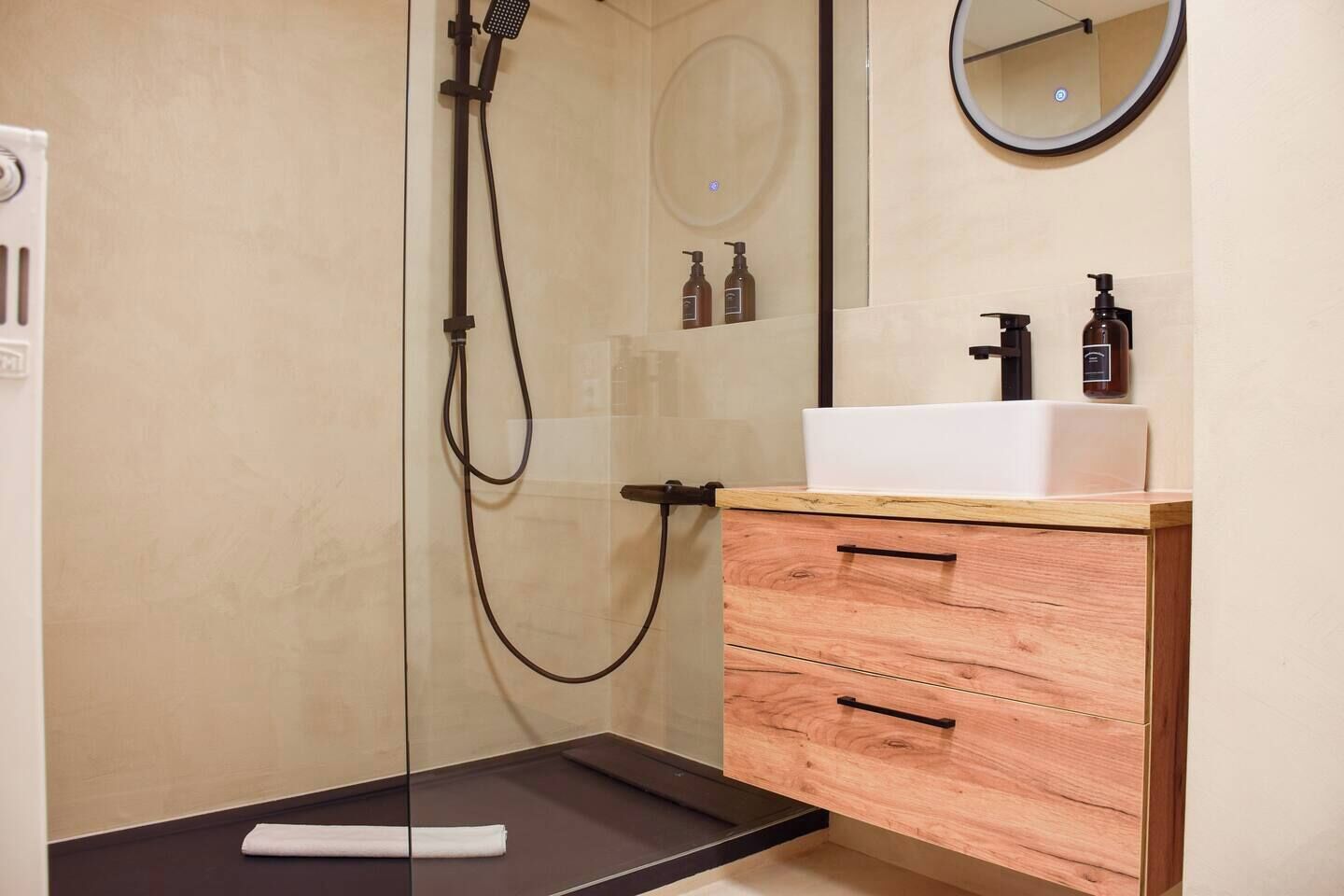 A modern bathroom with a glass shower enclosure, a wooden vanity with a white sink, and a round mirror.