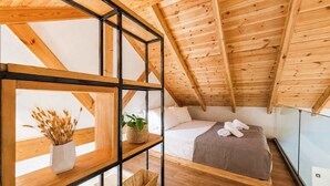 Ein Schlafzimmer mit schrägverlaufender Decke, Holzbalken, einem Bett mit weißen Leinen, einem Nachttisch mit Pflanze und einem Fenster.