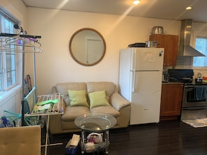 A compact living space with a sofa, a glass coffee table, a kitchen area including a refrigerator and stove, and a laundry rack.