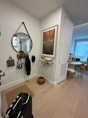 A room with a round mirror, a framed artwork, a small shelf with decorative items, a bench, and a suitcase. - Luxury DUMBO 1BR Spa + Gym Rooftop+Working Space (Brooklyn, NY)