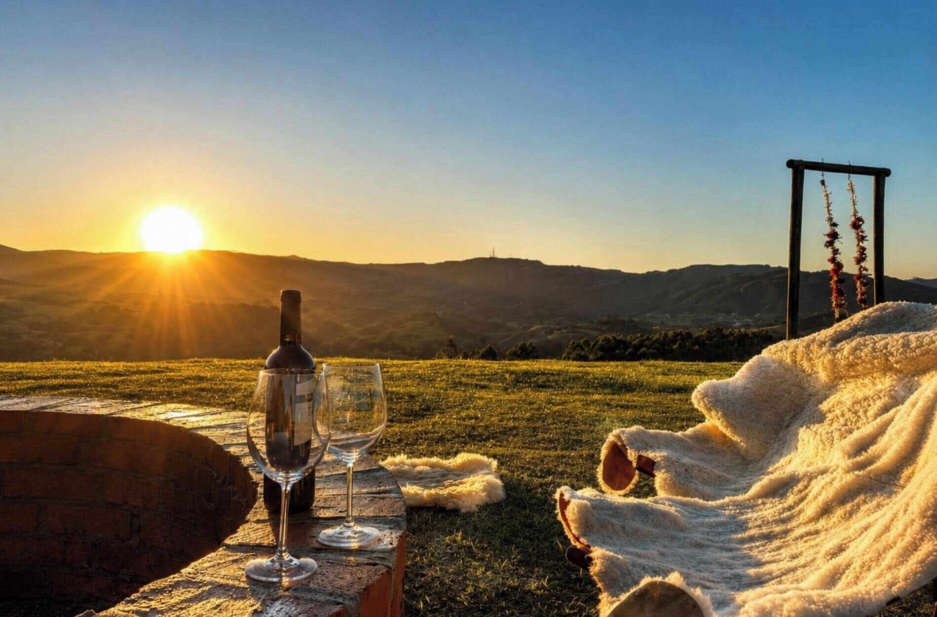 Ein Picknicktisch mit Flasche und Gläsern, eine Decke und Blick auf einen Sonnenuntergang über Hügeln.