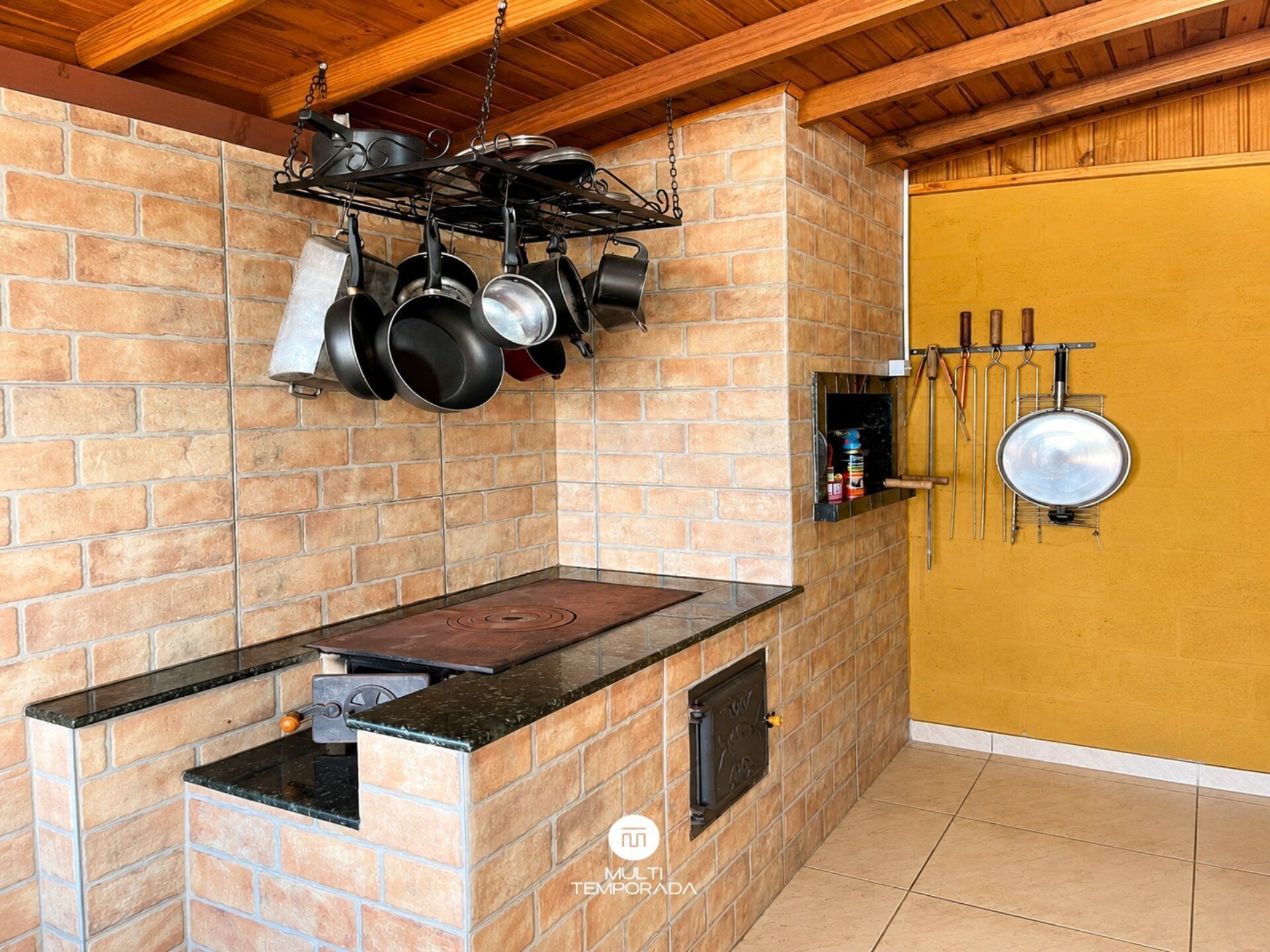 Cozinha com parede de tijolos, fogão, panelas penduradas e porta com parede amarela.