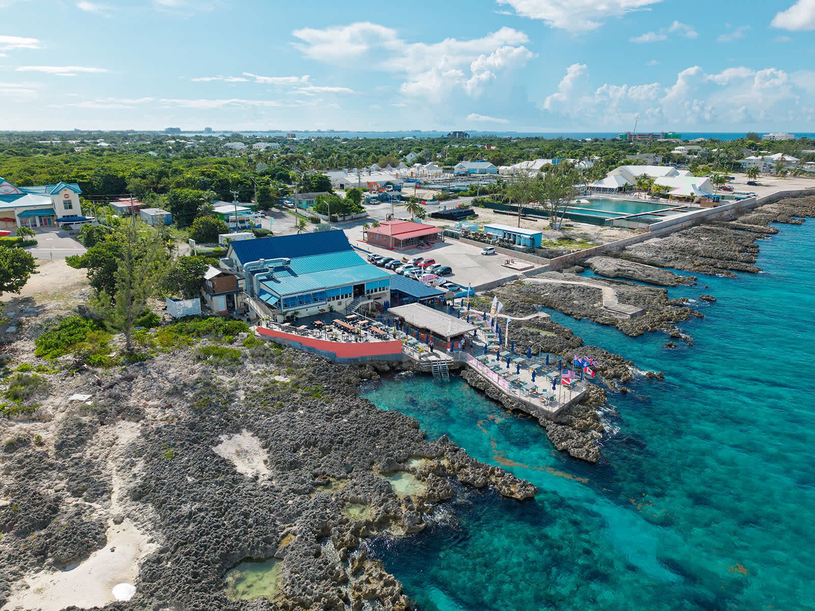 An aerial view of a coastal area with a prominent building with a blue roof, a pier extending into the turquoise sea, and surrounding greenery.