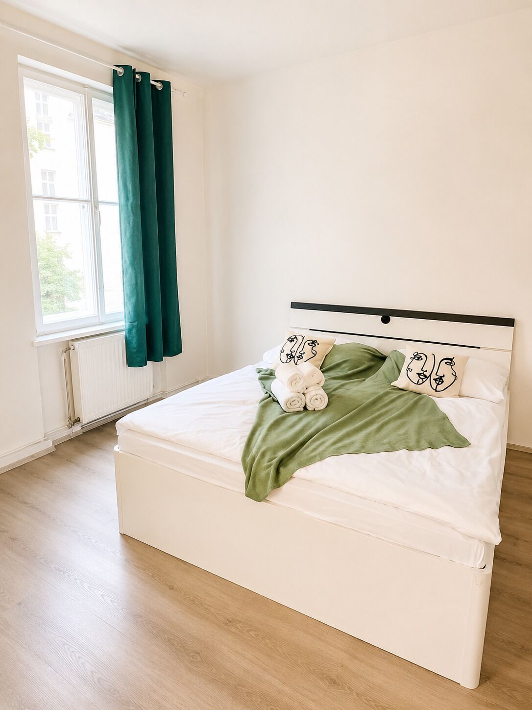 Un lit bien fait, avec une couverture verte et deux oreillers, une fenêtre avec des rideaux et un radiateur.