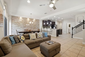 A modern living room with a sectional sofa, ottoman, and a dining area with a brick accent wall and pendant lighting. - Enchanted Pines Retreat!  Near Disney with Resort Pool (Davenport)