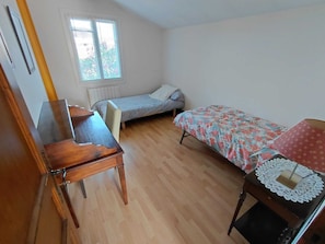 Ein kleiner, ordentlicher Raum mit Schreibtisch aus Holz, einem Einzelbett mit blumigem Überwurf, einem Fenster mit Jalousien und einer Heizung.