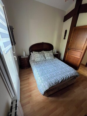 A bedroom with a wooden bed, a nightstand, a dresser, and a window with blinds. - Basque-style house with a 300 m2 garden, a sunny veranda, and a plancha (Anglet)