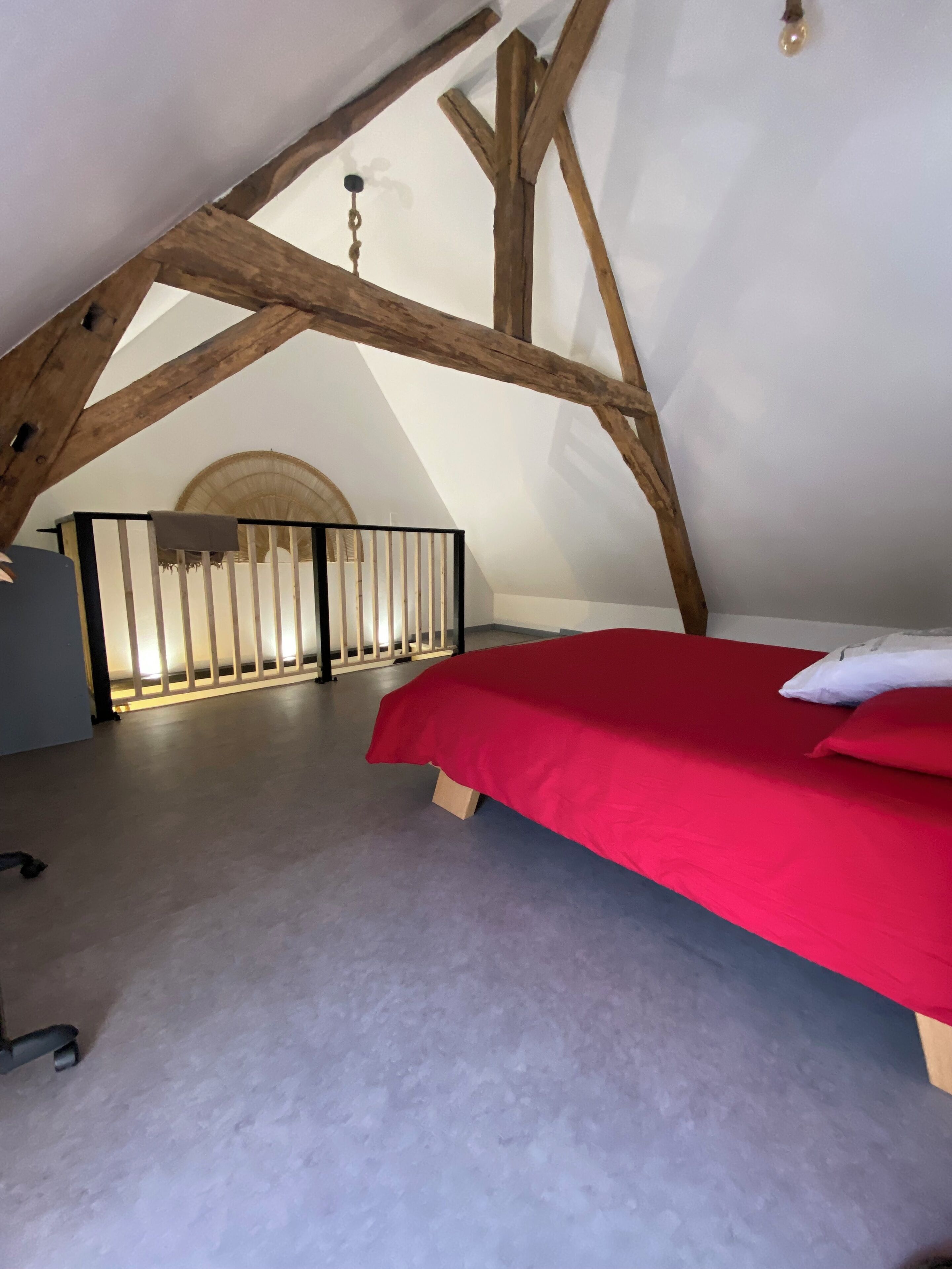 Una camera da letto mansardata con soffitto spiovente, travi in legno, un letto rosso e una ringhiera bianca.