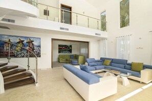 Un vaste espace de vie comprenant un escalier, un canapé d’angle bleu, un fauteuil vert et un tableau représentant des femmes portant des chapeaux.