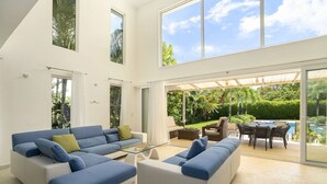 Ein großzügiger Wohnbereich mit großem Fenster, einer Sofagarnitur, einem Glastisch und Blick auf Pool und Garten.
