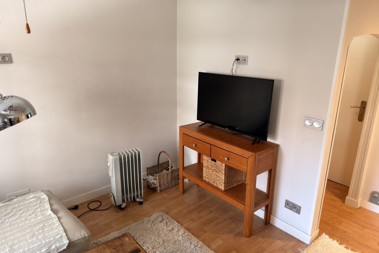 A room with a bed, a wooden TV stand with a flat-screen TV, a radiator, and a basket.