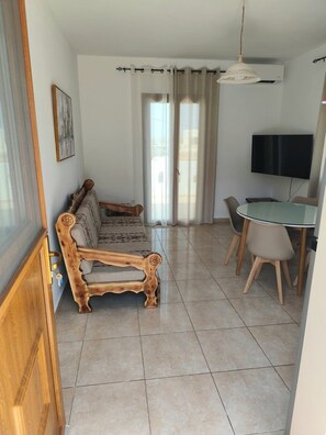A living room with a wooden sofa, a dining table with chairs, a TV, and a large window with curtains. - Naxos Chora Next (Naxos)