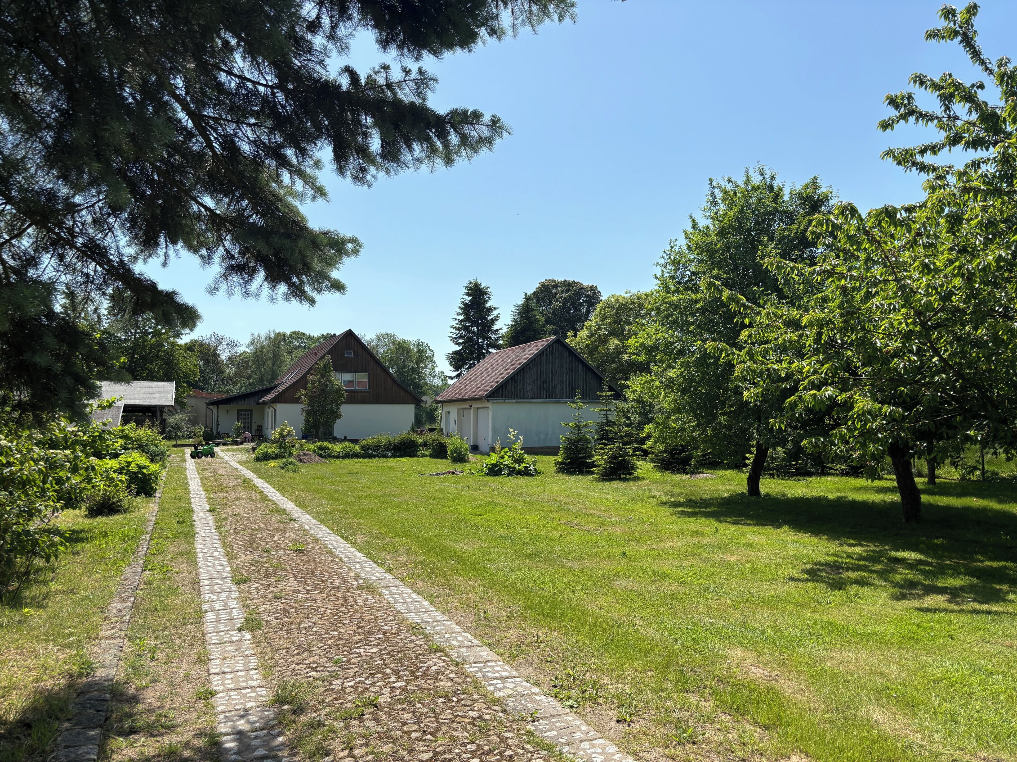 A rural setting with a paved path leading to a row of buildings, surrounded by greenery and trees.