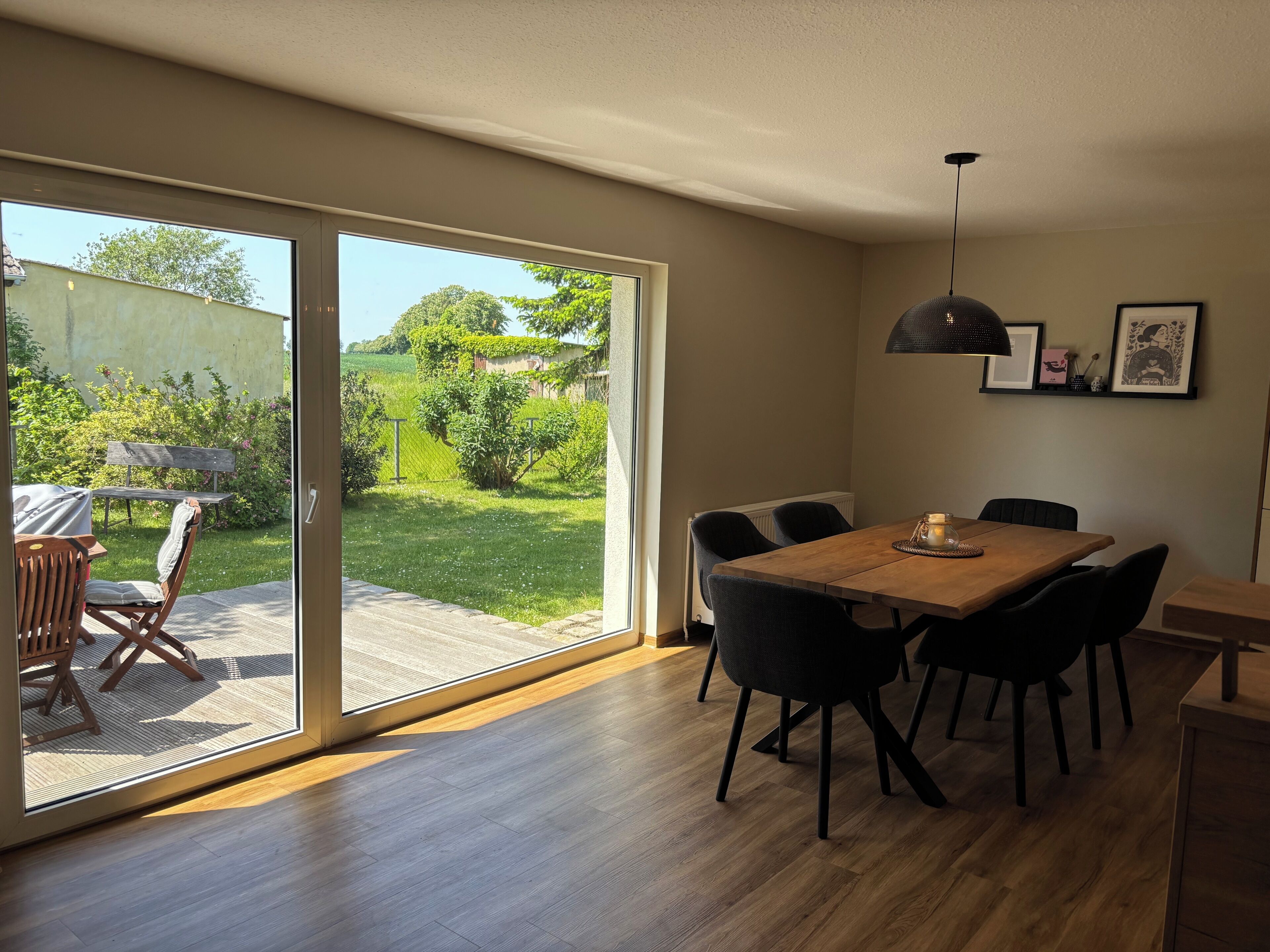 A dining area with a wooden table and chairs, a pendant light, and a view through a sliding glass door to a garden.