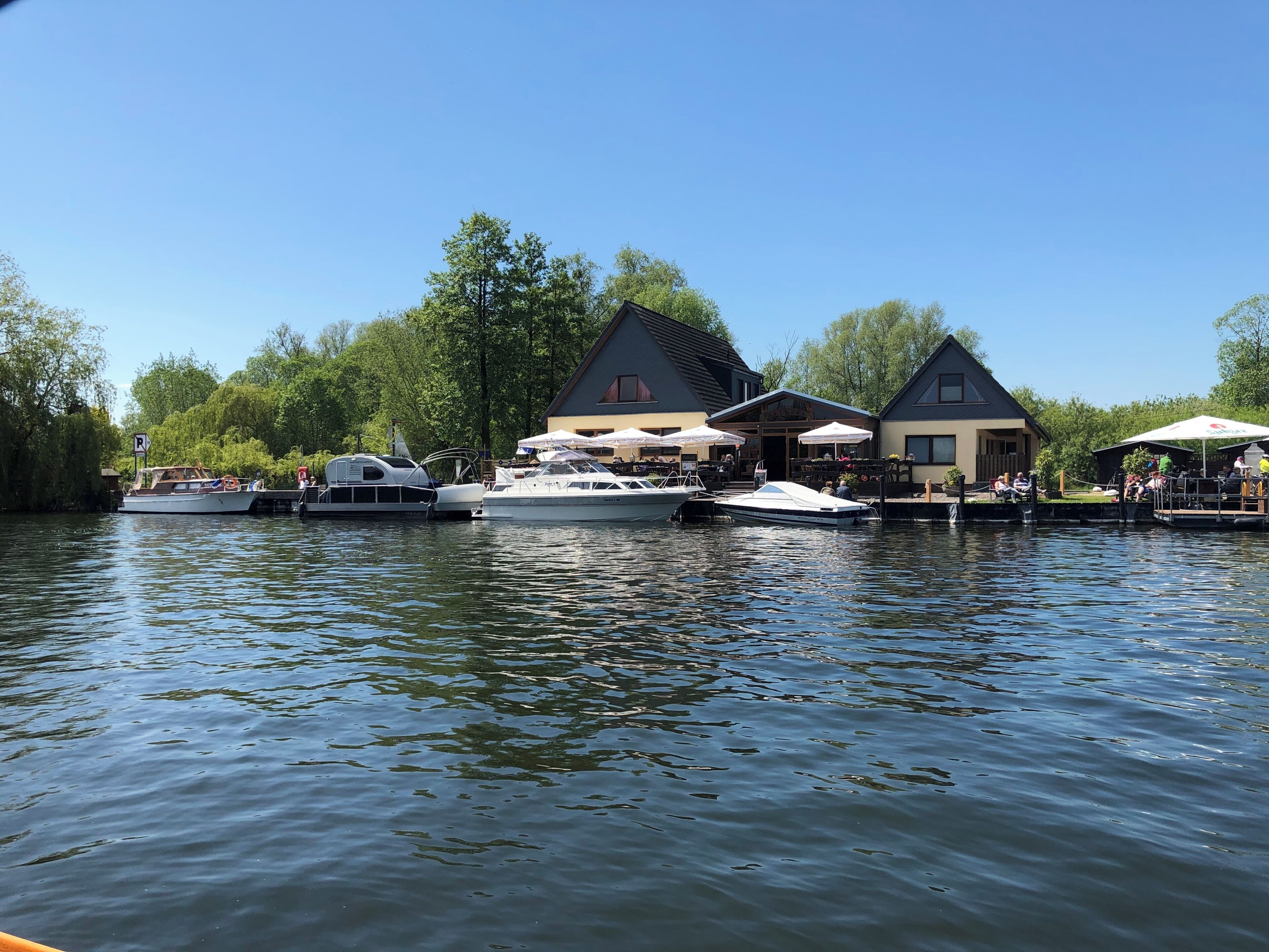 A riverside dock with boats, a restaurant with outdoor seating, and a building with a sloped roof.
