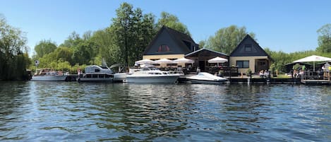 Un quai au bord de la rivière où sont amarrés des bateaux, un restaurant avec une terrasse, et un bâtiment doté d’un toit en pente.