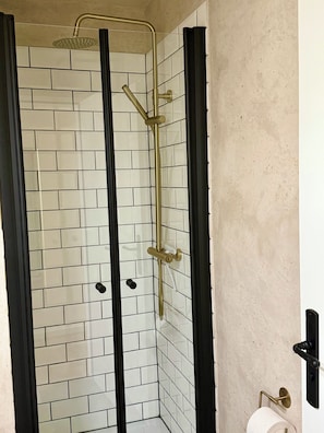 Um chuveiro envidraçado com chuveiro e painel de controle dourados, paredes azulejadas brancas e moldura de porta preta.