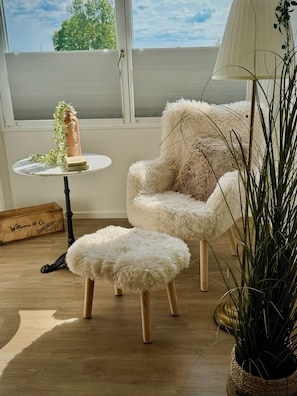 A room with a large window, a fluffy chair, a small round ottoman, a table with a vase and book, and a wicker plant.