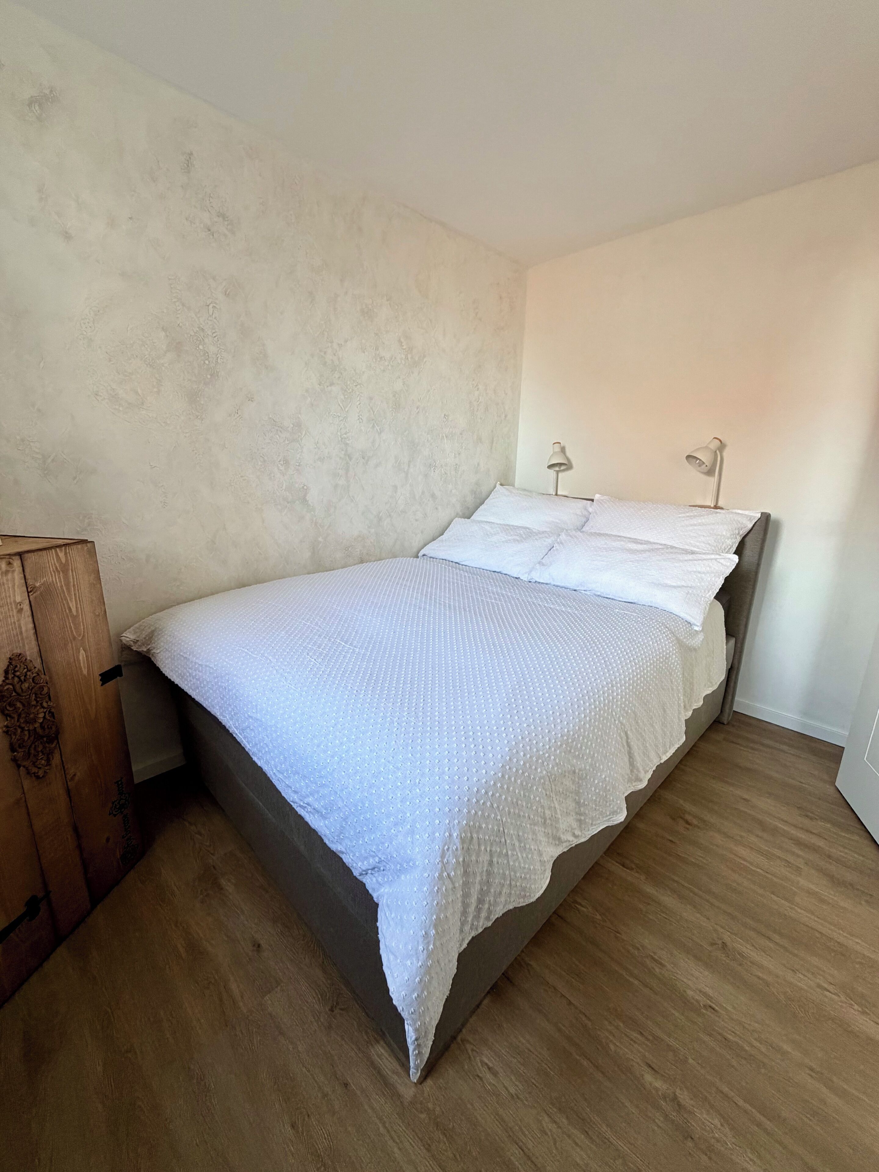 Uma cama de solteiro com um cobre-leito branco e almofadas, uma mesinha de cabeceira de madeira e duas luminárias.