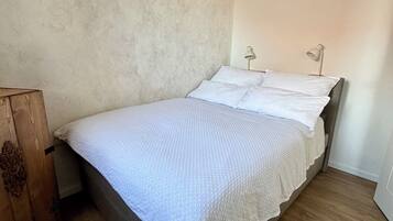 Uma cama de solteiro com um cobre-leito branco e almofadas, uma mesinha de cabeceira de madeira e duas luminárias.