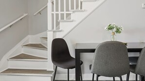 Uma escadaria com grades brancas e degraus de madeira, uma mesa de jantar com cadeiras e um vaso com flores.