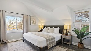 Quarto com cama, mesas de cabeceira, um vaso de planta e janela com cortinas.