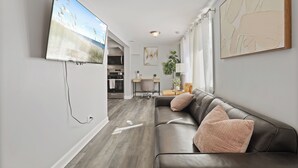 Uma sala de estar moderna com sofá de couro, televisão de tela plana na parede, área de jantar com mesa e cadeiras, e cozinha ao fundo.