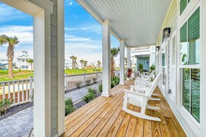Eine Holzterrasse mit weißen Stühlen, einem weißen Zaun und Blick auf ein Haus und Palmen.