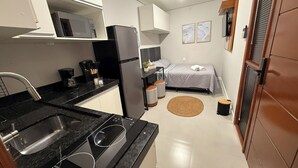 A compact kitchen with a sink, stove, and microwave, a small dining area, a bed with a gray bedspread, and a round rug. - LOFT127 - 02 (Itajaí)