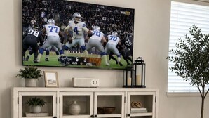Ein Wohnzimmer mit Fernseher, der ein Fußballspiel zeigt, einem weißen Schrank mit Glastüren, einem Holztisch und einer Topfpflanze.