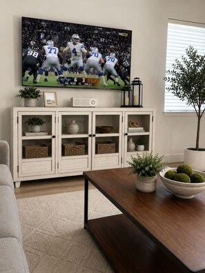 Ein Wohnzimmer mit Fernseher, der ein Fußballspiel zeigt, einem weißen Schrank mit Glastüren, einem Holztisch und einer Topfpflanze.