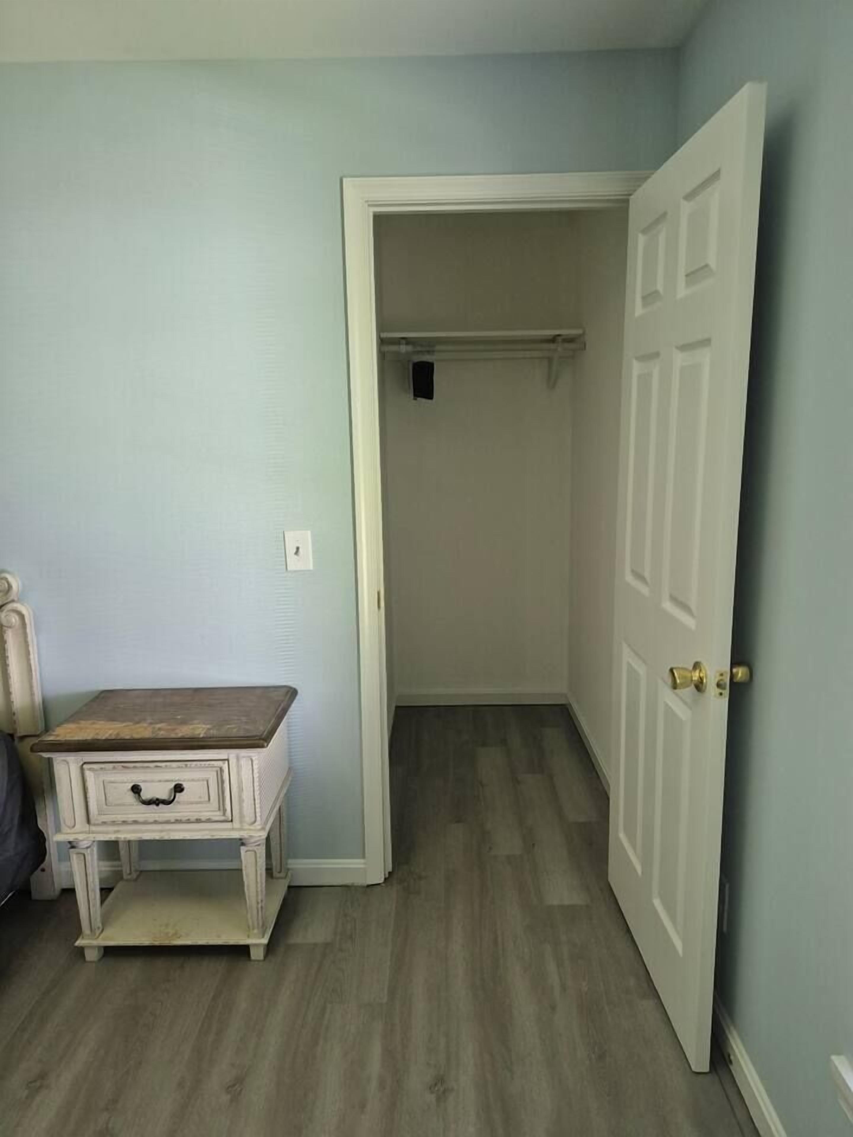 Um quarto com piso de madeira, uma porta branca e uma pequena mesa branca com gavetas.
