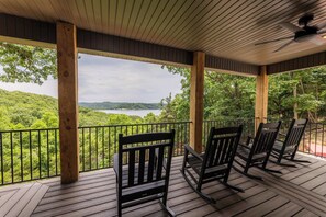 A covered deck with wooden railings, rocking chairs, and a view of a lake surrounded by trees. - Oak Ridge on Beaver Lake, 5BR Lakeview, Sleeps 13 (Garfield)