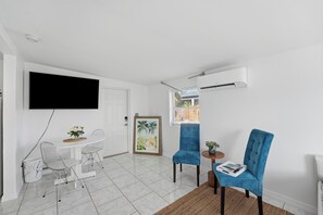 Ein modernes Wohnzimmer mit Flachbildfernseher, einem Esstisch mit Stühlen, einem blauen Sessel und einem kleinen Beistelltisch mit einer Zeitschrift.