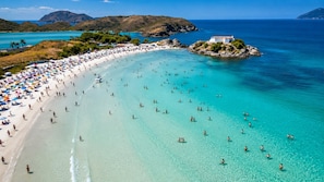 A beach with clear turquoise water, a white sandy shore, and a small white building on a rocky outcrop.