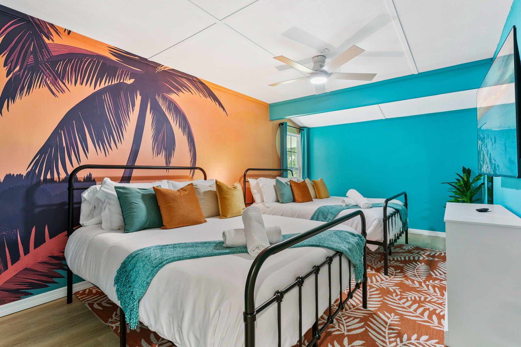 A room with two beds, a ceiling fan, a large mural, and a desk.
