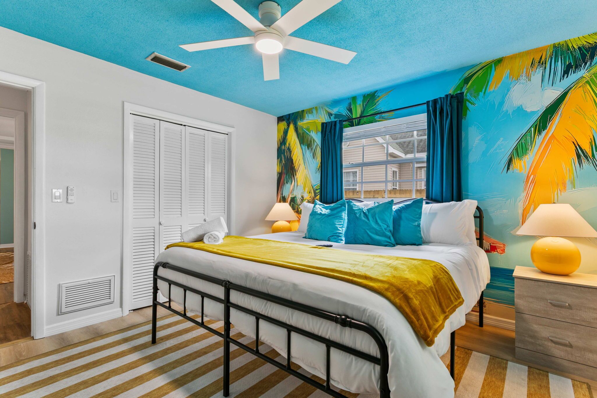 A bedroom with a bed, a ceiling fan, a window with curtains, a nightstand, and a wall mural.