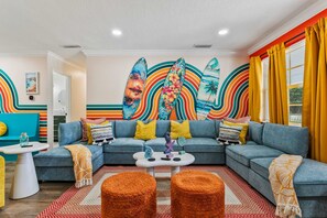 A modern living room with a sectional sofa, colorful artwork, and a bright window with yellow curtains. - The Rainbow Lagoon - Pickleball, Heated Pool, Golf (Bradenton)