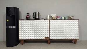 Et køkkenområde med et sort køleskab, et hvidt skab med geometriske mønstre, en kaffemaskine og diverse køkkenartikler på bordet.