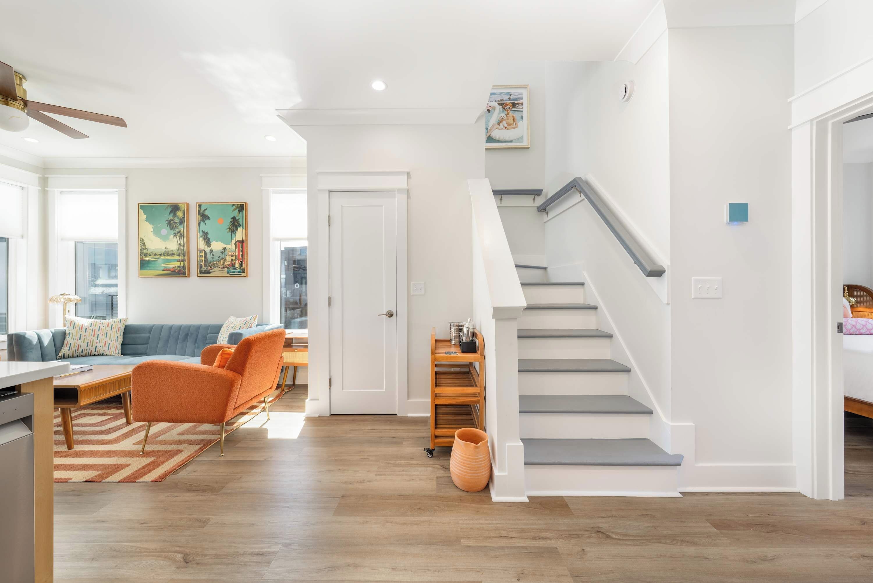 Een ruime woonkamer met een trap, houten vloeren en een eethoek met een blauwe bank en oranje stoelen.