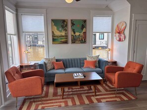 Ein Wohnzimmer mit grauem Sofa, orangenen Sesseln, einem Holztisch und zwei Bildern an der Wand.