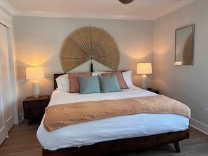 Cama bem arrumada com cabeceira, duas mesas de cabeceira com abajures e um ventilador de parede grande.