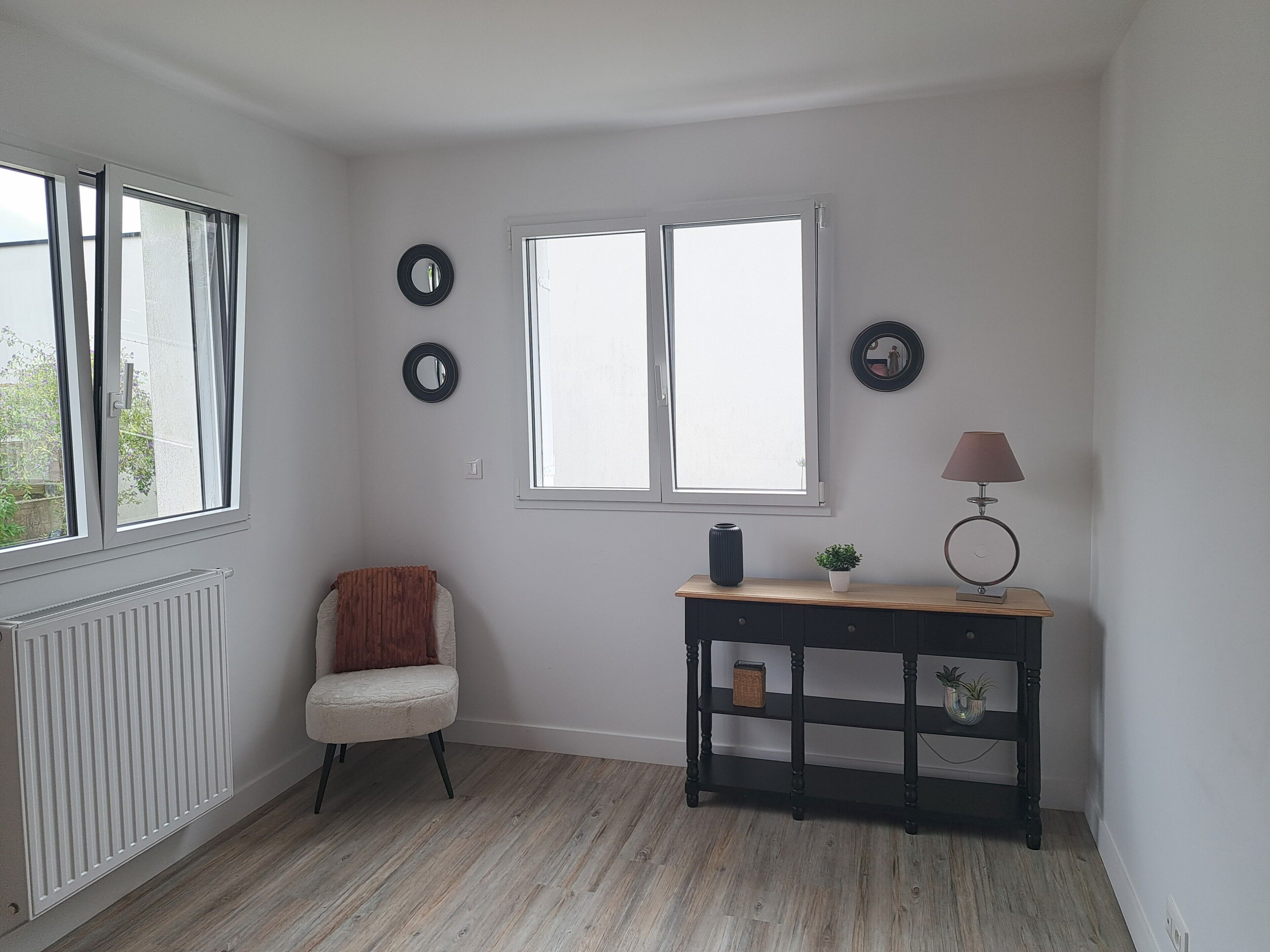 A room with a radiator, a chair, a table with a lamp, and two windows.